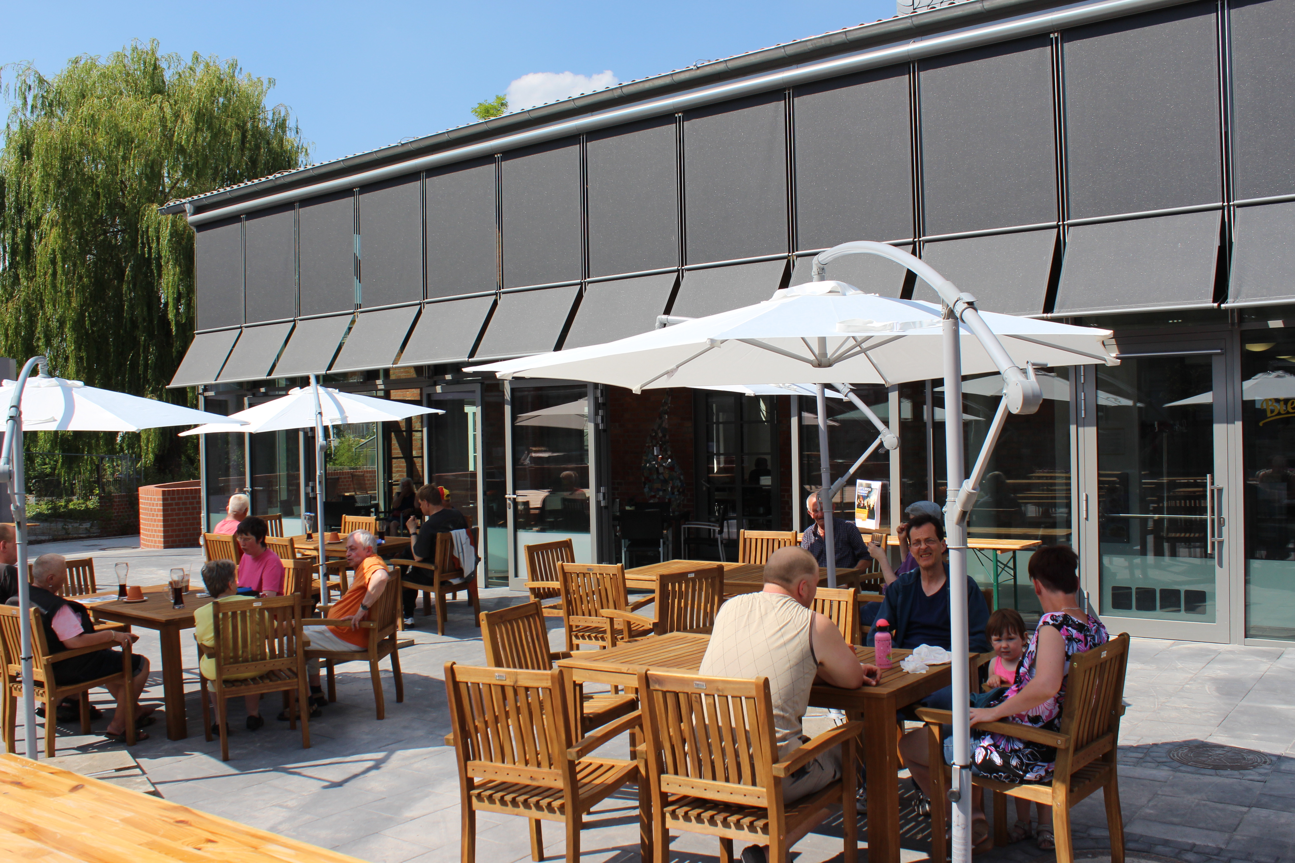 Gebäude des "Biermanskis" von Außen. Sitzgruppen mit weißen Sonnenschirmen und Menschen, die Essen und das Wetter genießen