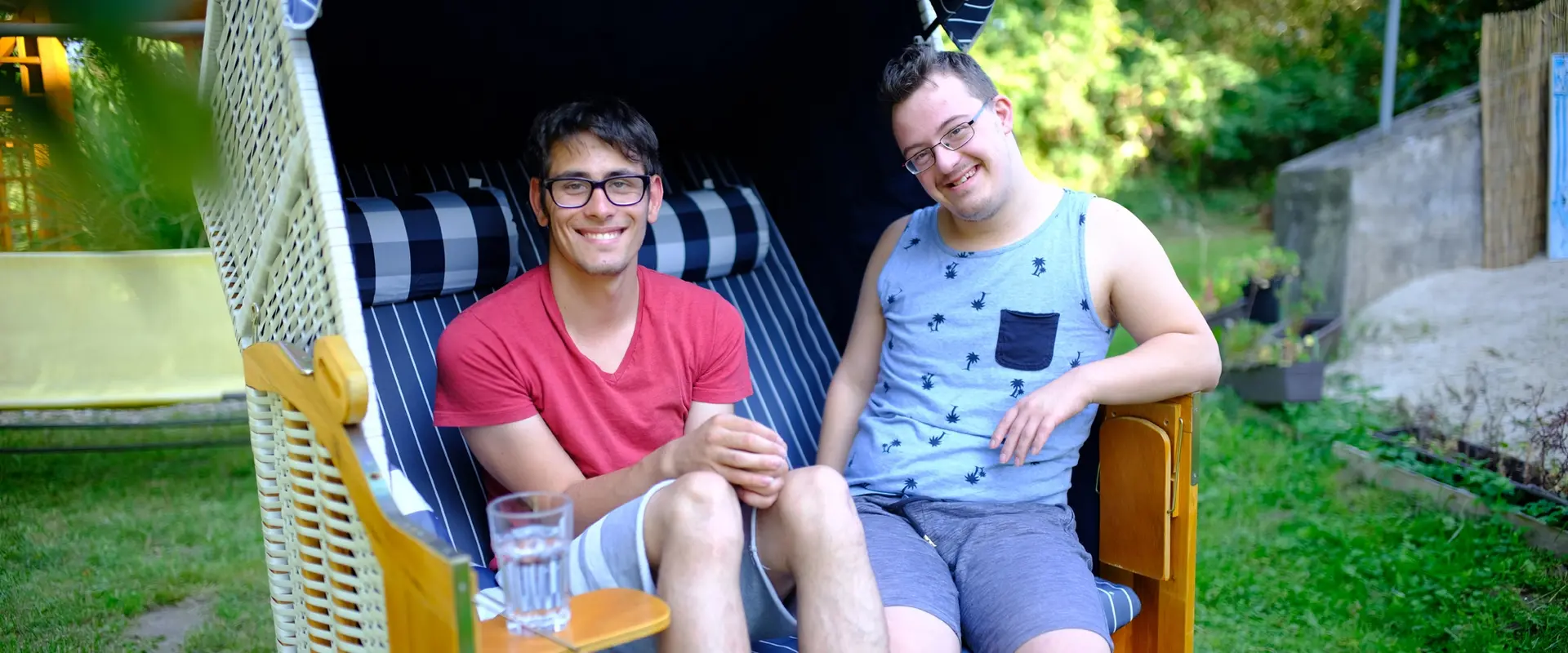 Zwei junge Männer mit Behinderungen sitzen in einem Strandkorb und lachen in die Kamera