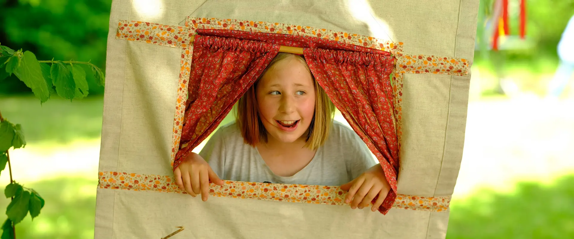Mädchen schaut durch einen hellen Spielvorhand, in dem ein Fenster mit Roten Gardinen ist.