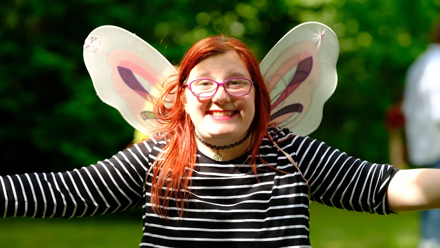 Mädchen lacht mit ausgebreiteten Armen in die Kamera, sie trägt Schmetterlingsflügel
