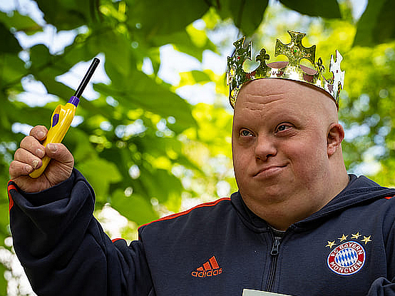 Mann mit Downsyndrom trägt eine Krone, hält ein Feuerzeug hoch und ein Buch unter dem Arm. Er ist Schauspieler im Theater Endlich