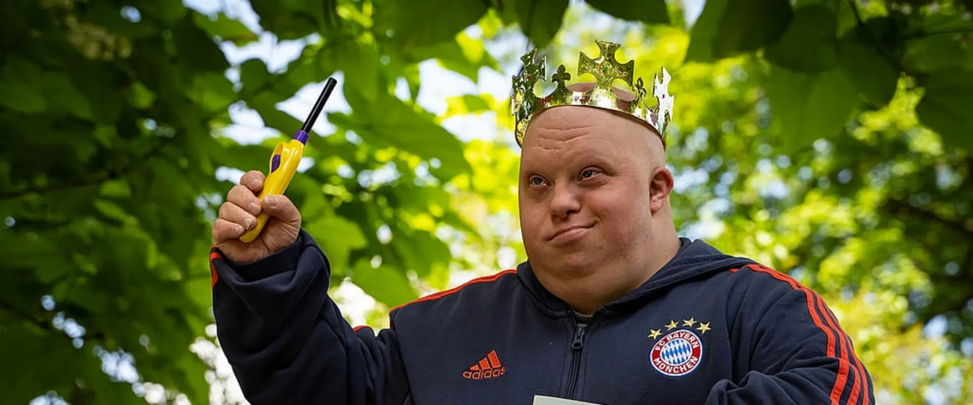Mann mit Downsyndrom trägt eine Krone, hält ein Feuerzeug hoch und ein Buch unter dem Arm. Er ist Schauspieler im Theater Endlich